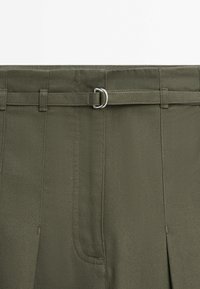 Gros plan d’un pantalon vert olive avec une ceinture en tissu passée dans une boucle double anneaux argentée à la taille.