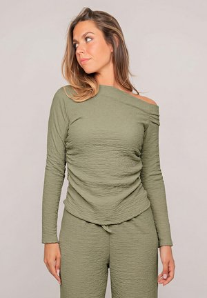 Femme aux cheveux mi-longs portant un haut texturé vert olive à épaules dénudées avec manches longues et un pantalon assorti, regardant vers la gauche.