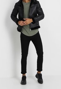 Schwarze Lederjacke mit Pelzbesatz, olivgrünes Hemd, schwarze schlanke Hose und schwarze Turnschuhe. Einfaches Design mit Reißverschluss.