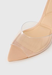 Mule beige à bout pointu avec une large sangle transparente à l'avant et un coussin rembourré avec des coutures géométriques sur la semelle intérieure.