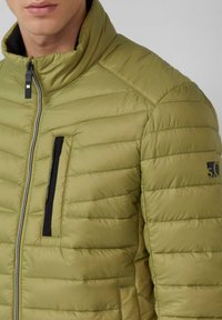 Oliwkowa pikowana kurtka puchowa z zamkiem z przodu, z poziomymi przeszyciami, stójką oraz zapinaną na zamek kieszenią na piersi z czarnymi akcentami.