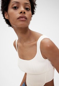 Ribbet hvid croptop med brede stropper og firkantet udskæring. Glat tekstur og tætsiddende design. Modellen bærer denimnederdel.