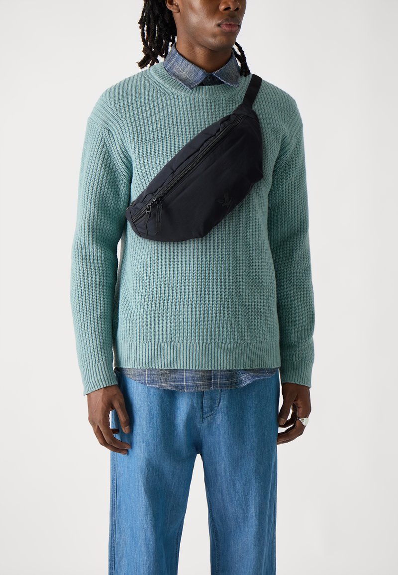 Una cintura nera in tessuto indossata sopra un maglione a maglia verde menta. Il maglione presenta una texture a coste e maniche lunghe. Pantaloni in denim blu completano l'outfit.