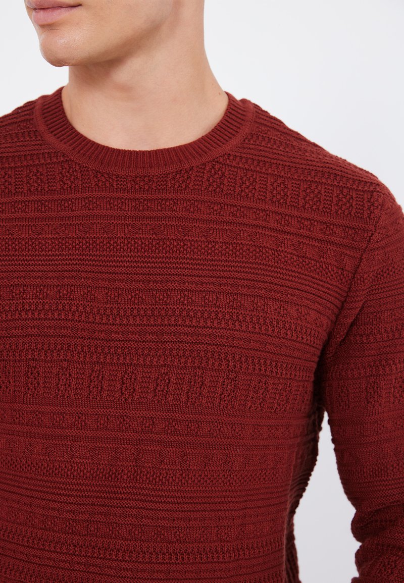 Maglione lavorato a maglia color mogano con scollatura rotonda, caratterizzato da modelli testurizzati e orlo a costine. Il tessuto mostra righe e intrecci alternati.