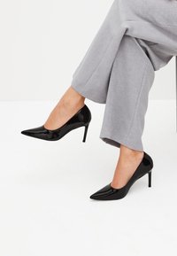 Zapatos de tacón alto de charol negro con punta afilada y diseño elegante; combinados con pantalones de pierna ancha de color gris claro.
