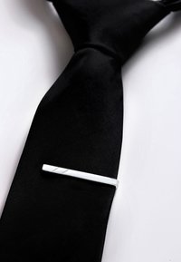 Hopeinen solmionpidike, jossa on minimalistinen muotoilu, sileä pinta ja vinot kaiverrukset, asetettuna mustan silkkinauhan päälle.