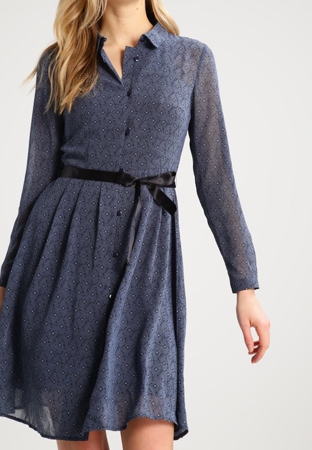 Femme portant une robe bleue à manches longues à motifs avec des boutons et une ceinture ruban noire nouée à la taille.