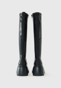 Pair of tall black leather boots with chunky heels viewed from the back, featuring vertical black straps and side zippers.