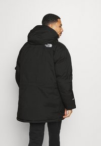 Zwarte geïsoleerde jas met een capuchon, voorzien van een logo op de schouder, elastische manchetten en een ontspannen pasvorm. Gladde textuur en ontwerp tot op de knie.