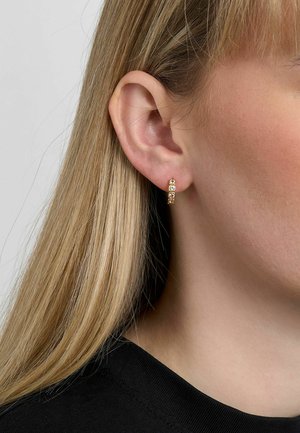 Gold hoop earring with three small gemstones worn on the left ear of a person with long blonde hair and black top.