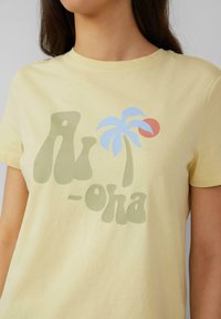 Lichtgele katoenen t-shirt met een grote afbeelding met groene tekst "Aloha," een blauwe palmboom en een rood zonnetje. Eenvoudig crew neck ontwerp.