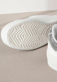 Adidași albi cu o talpă din cauciuc texturat, având un model în zigzag. Partea superioară este dintr-un material neted, cu un accent argintiu la călcâi.