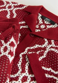Červený a biely pletený sveter so vzorovaným dizajnom, zapínaním na gombíky, golierom a štítkom s nápisom „NEXT“ vo vnútri výstrihu.