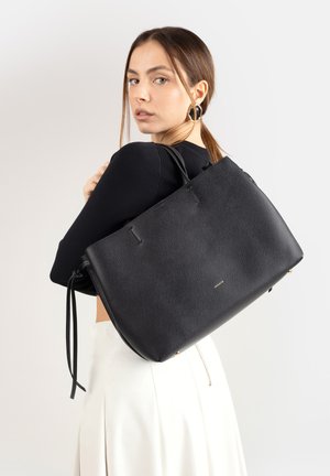 Donna con capelli castano chiaro, indossa un top nero e una gonna bianca, porta una grande borsa tote in pelle nera sulla spalla.