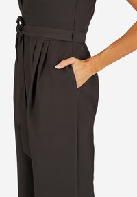 Zwarte jumpsuit met een aansluitende bovenkant en een geknoopt riempje, plooien bij de taille en zijzakken. Gladde stof met een matte afwerking.