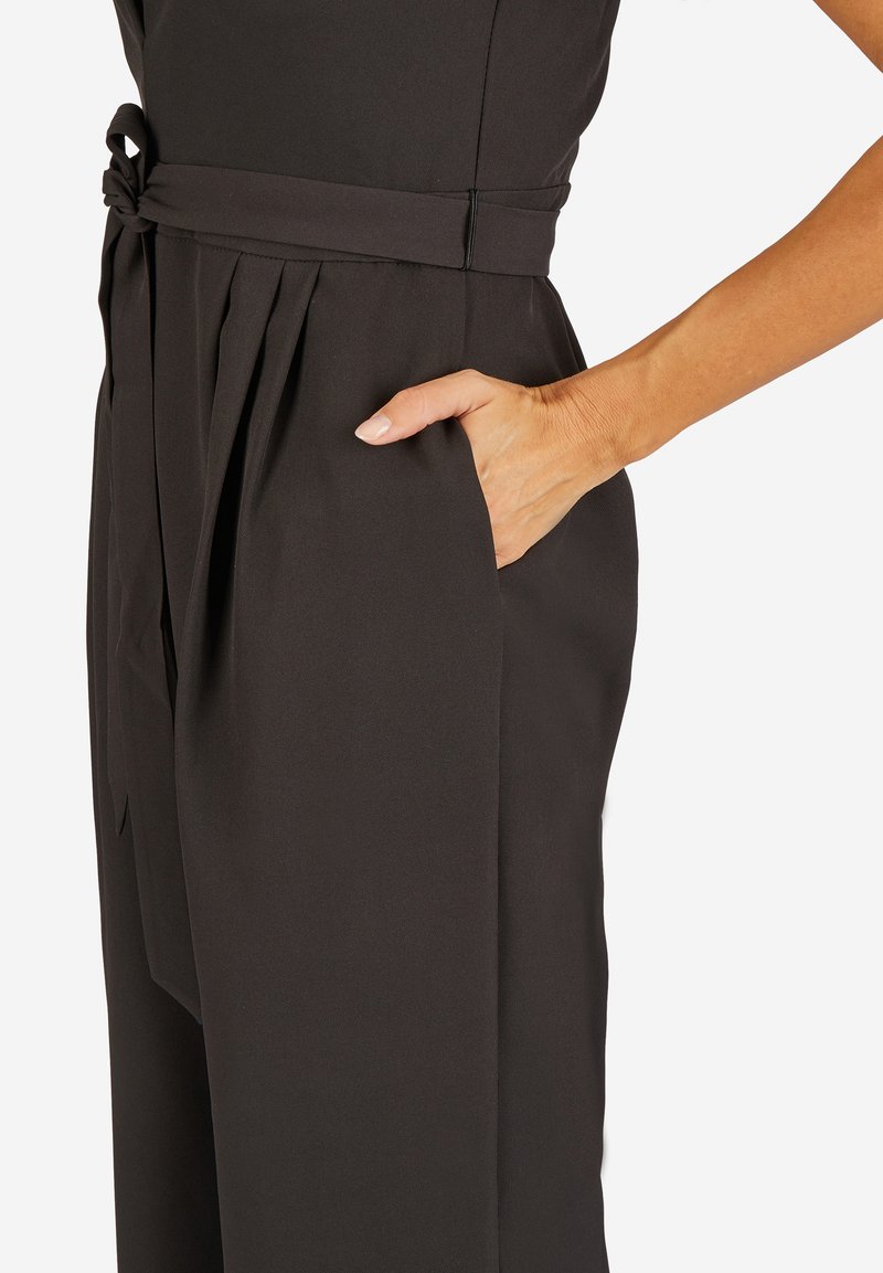Schwarzer Jumpsuit mit einem taillierten Oberteil, einem geknoteten Gürtel, Falten an der Taille und seitlichen Taschen. Glatter Stoff mit matter Oberfläche.