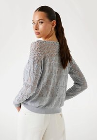 Light gray knitted sweater featuring an openwork design with diamond patterns, ribbed cuffs, and hem. Model faces slightly away from the camera.