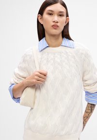 Krémový svetr s pleteným vzorom a vystúpenými vzormi, položený na modro-bielom pruhovanom košeľovom tričku, doplnený svetlo béžovou taškou.