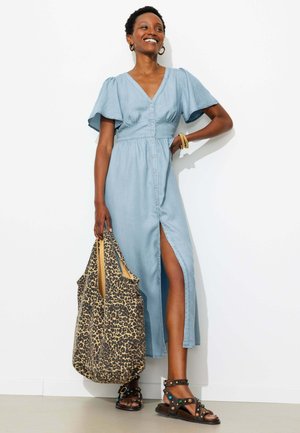 Femme en robe bleu clair boutonnée avec une fente latérale, tenant un grand sac imprimé léopard, portant des sandales noires cloutées et des boucles d'oreilles créoles dorées.