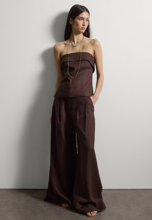 Joven mujer con top marrón sin tirantes y pantalones anchos a juego, de pie con una mano en el bolsillo frente a una pared lisa y clara.