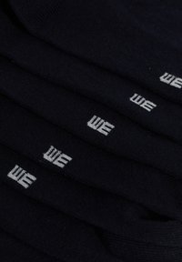 Črne nogavice v kupu, izdelane iz mehkega materiala, z belim vezenim logotipom "WE" na vsakem paru. Gladka tekstura, standardna dolžina do gležnjev.