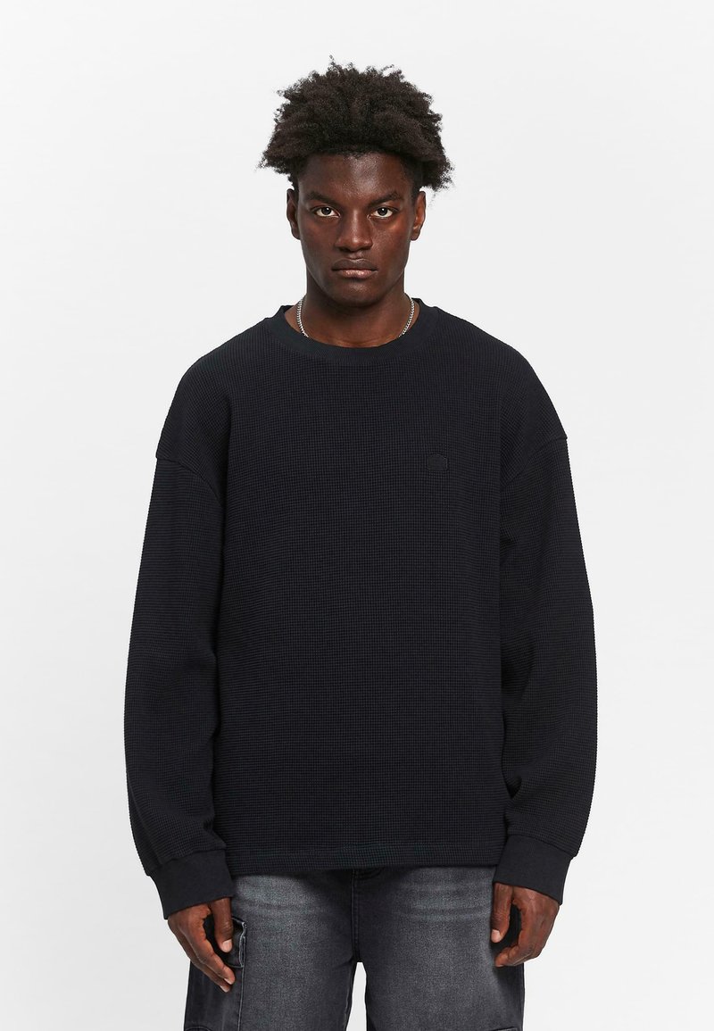 Svart oversized hettegenser med en teksturert vaffelmønster og ribbestrikkede mansjetter. Har en subtil logo på brystet.