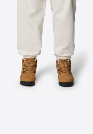 Brune snørebåndsstøvler med overdel i ruskinn, sorte gummisåler og metallegale, kombinert med hvite bukser med elastisk ribb.