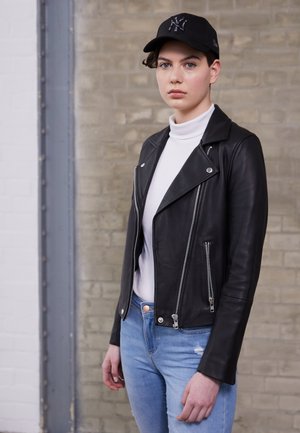 Schwarze Lederbikerjacke mit silbernen Reißverschlüssen, über einem weißen Pullover mit Rollkragen getragen. Hellblaue, ausgebleichte Jeans und eine schwarze Mütze runden den Look ab.