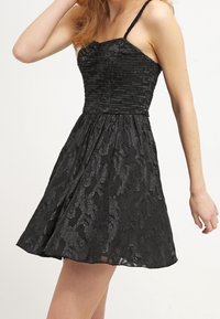 Robe noire avec des bretelles spaghetti, corsage froncé et jupe évasée. Présente un motif texturé avec un subtil brillant. Tissu léger.