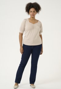 Beige topp med kort ärm och fyrkantig halsringning samt rynkdetalj, ihopparad med mörkblå slim-fit jeans och beige lägenheter. Enkel, slät tyg.