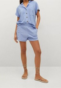 Camisa de manga corta azul clara con cuello, tres botones y un bolsillo en el pecho, combinada con pantalones cortos a juego; sandalias planas con ataduras en el tobillo.