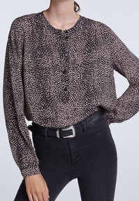 Leopardenmuster-Bluse aus leichtem Stoff, mit langen Ärmeln, runder Halslinie und Knopfdetails an der Vorderseite. Getragen mit schwarzen Hosen.