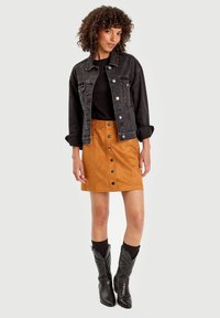 Veste en denim noir, chemise noire et mini-jupe en suède beige avec des boutons sur le devant. Des bottes de cow-boy noires complètent la tenue.