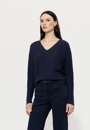 Marineblauer V-Ausschnitt-Pullover mit langen Ärmeln, Strickstruktur und lockerer Passform, kombiniert mit dunklen Jeans aus Denim mit Fronttaschen.