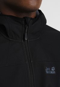 Giacca softshell nera con colletto alto, zip frontale e logo sul lato sinistro. Presenta una texture liscia e dettagli sulle cuciture.