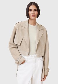 Beige Wildleder-Cropped-Jacke mit breitem Kragen, asymmetrischem Design und silbernen Hardware-Details. Kombiniert mit einem hellen Oberteil und weißen Hosen.