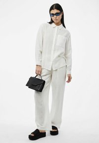 Camisa blanca de botones y pantalones a juego, de corte holgado y en tejido ligero. Bolso negro estructurado y sandalias de plataforma. Gafas de sol azules.