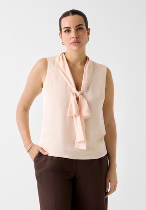 Kobieta z ciemnymi włosami zebranymi do tyłu, ubrana w bladoróżową bezrękawnikową bluzkę z wiązanym szalikiem na szyi i ciemnobrązowe spodnie, stojąca na neutralnym tle.
