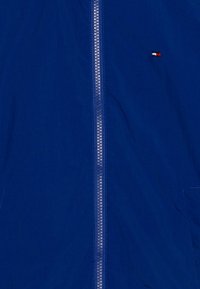 Niebieska kurtka z przednim zamkiem błyskawicznym i małym logo w kolorach czerwonym, białym i niebieskim. Lekka tkanina o gładkiej fakturze.