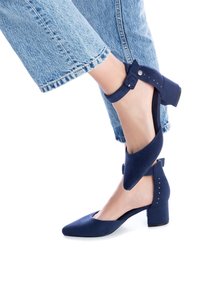 Fötter i marinblå låg blockklacksskor med ankelsnören, tillsammans med ljusblå denimjeans mot en vit bakgrund.