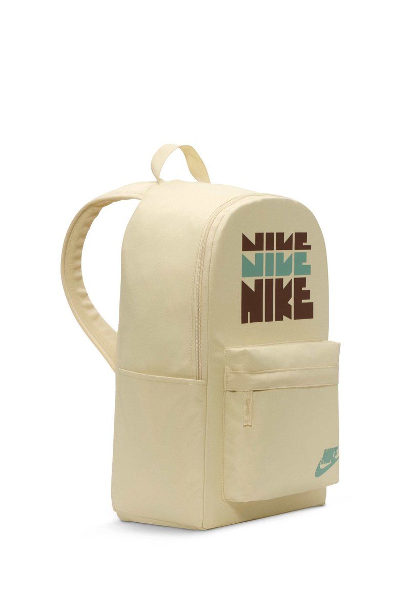 Mochila de color crema con forma redondeada, gran bolsillo frontal y el logo de "Nike" en marrón y turquesa en el lado. La textura de la tela es suave.
