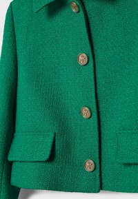 Zielona teksturowana kurtka z złotymi guzikami, kołnierzem i dwoma przednimi kieszeniami. Tkanina ma wzór tkany, dodając głębi jej wyglądowi.