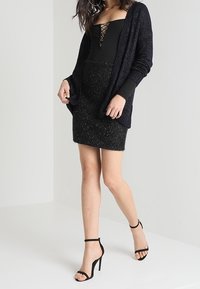 Černý dlouhý kardigan z texturovaného pleteného materiálu, doplněný těsnými černými šaty s vázáním na výstřihu a černými páskovými lodičkami.