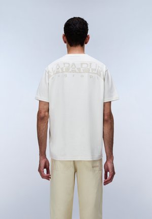 Homme vu de dos portant un T-shirt blanc manches courtes avec le texte "NAPAPIJRI" sur le haut du dos et un pantalon beige clair.