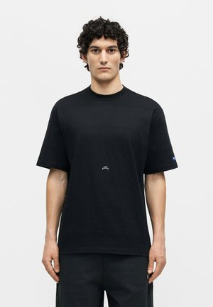 Νέος άνδρας με σγουρά μαλλιά που φοράει ένα απλό μαύρο κοντομάνικο μπλουζάκι και μαύρο παντελόνι, στέκεται μπροστά σε ανοιχτό φόντο.