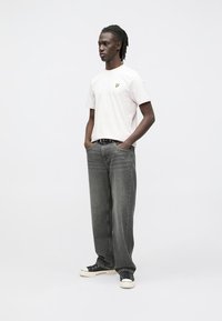 Lyserød t-shirt med korte ærmer og en gul logo på brystet, kombineret med løstsiddende mørkegrå jeans og sorte sneakers.