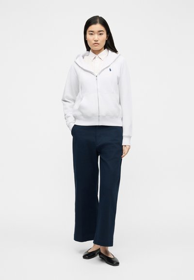 Giovane donna con felpa con zip bianca, camicia a righe con bottoni, pantaloni larghi blu scuro e ballerine nere in piedi davanti a uno sfondo semplice.