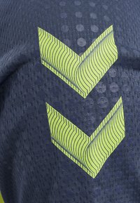 Twee gele chevrons met dunne zwarte lijnen op donkerblauw gestructureerd stof met cirkelpatronen.