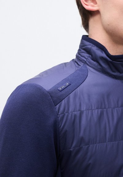 Άνδρας που φοράει σκούρο μπλε μπουφάν με καπιτονέ ύφασμα, πλεκτά μανίκια και γιακά, με μικρό λογότυπο "RLX" στην πλευρική ραφή στον ώμο.