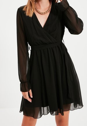 Frau trägt ein schwarzes, langärmliges Chiffon-Wickelkleid mit Taillenschärpe und einer goldfarbenen, massiven Kettenhalskette, die Hand in Kopfnähe erhoben.